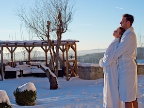 A couple embracing in a snowy outdoor setting with a wooden pergola and snow-covered trees. - Panoramahotel Grobauer (Spiegelau)