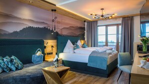 A modern hotel room with a large bed, a wooden headboard, a view of the city, and a cozy seating area. - Panoramahotel Grobauer (Spiegelau)