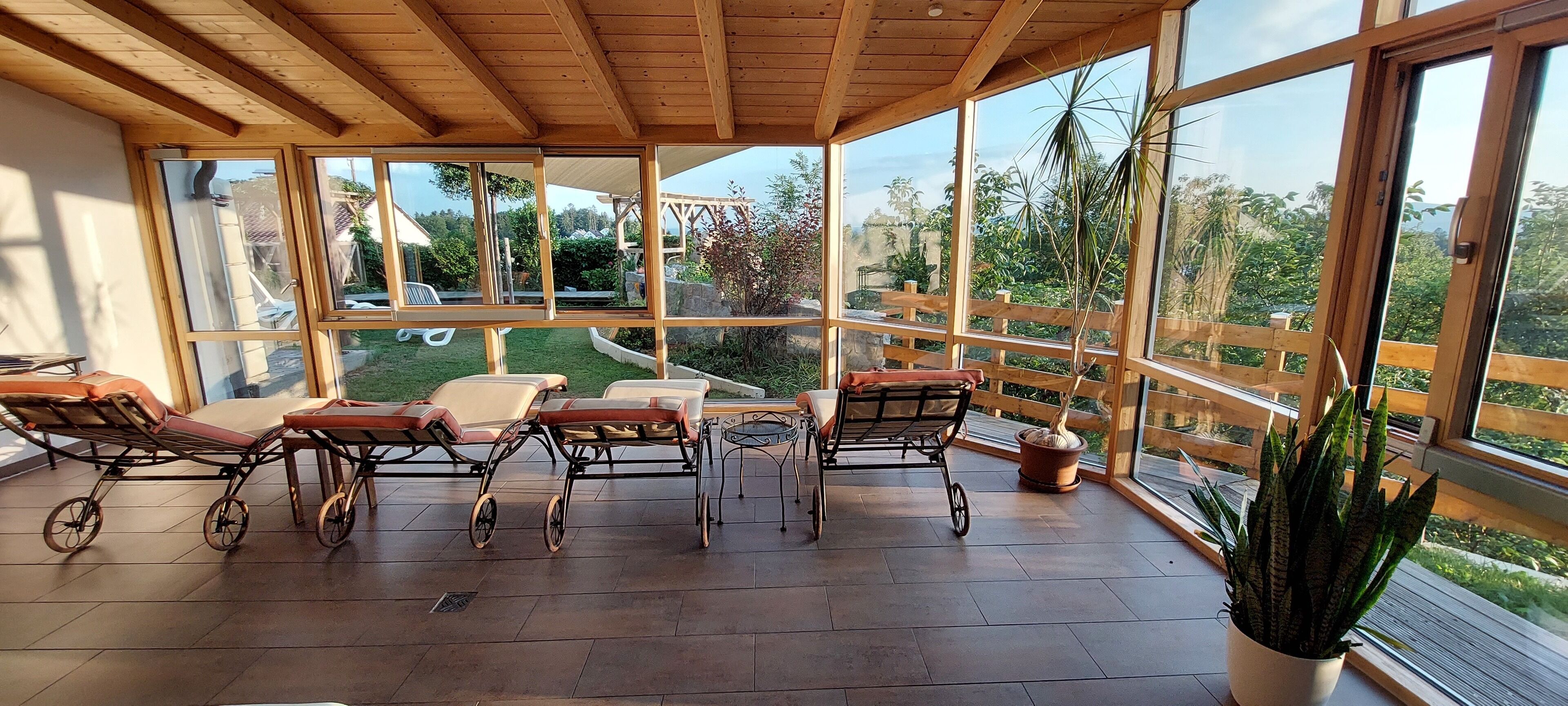 A covered outdoor area with wooden flooring, multiple chairs, a table, and large windows offering a view of the garden and surrounding area.