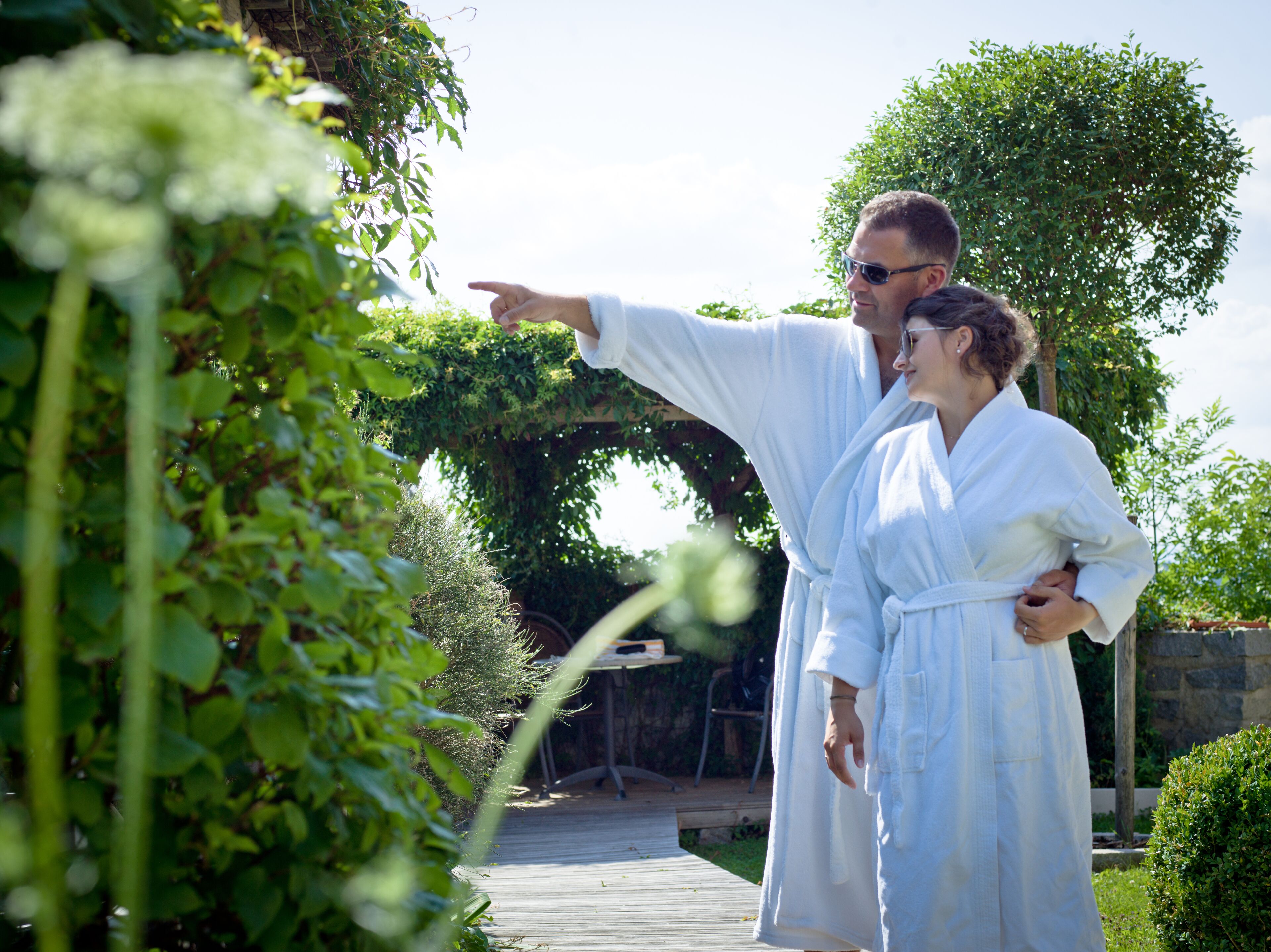 A couple in white robes standing on a wooden deck with greenery in the background.
