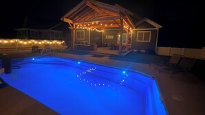 Uma piscina iluminada em azul, um espaço de estar coberto com luzes de fada e uma casa com deck de madeira.