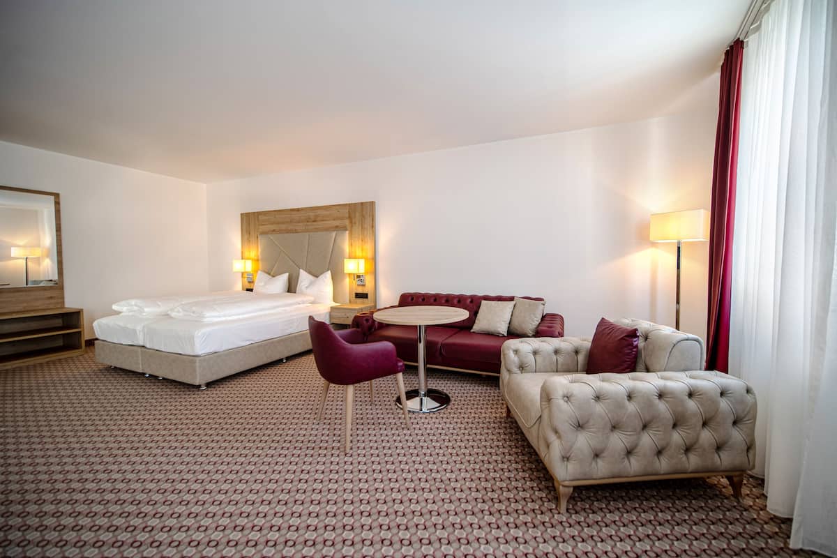 Una habitación de hotel con una cama, un sofá rojo, un sillón beige, una mesita redonda y una lámpara.