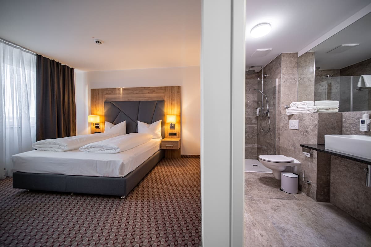 Una habitación de hotel con cama, lámparas de noche, baño con lavamanos, inodoro y ducha.