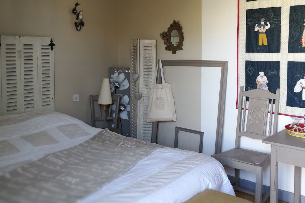 A bedroom with a bed, a desk, and a chair. There is a wall with a quilted panel and a mirror.
