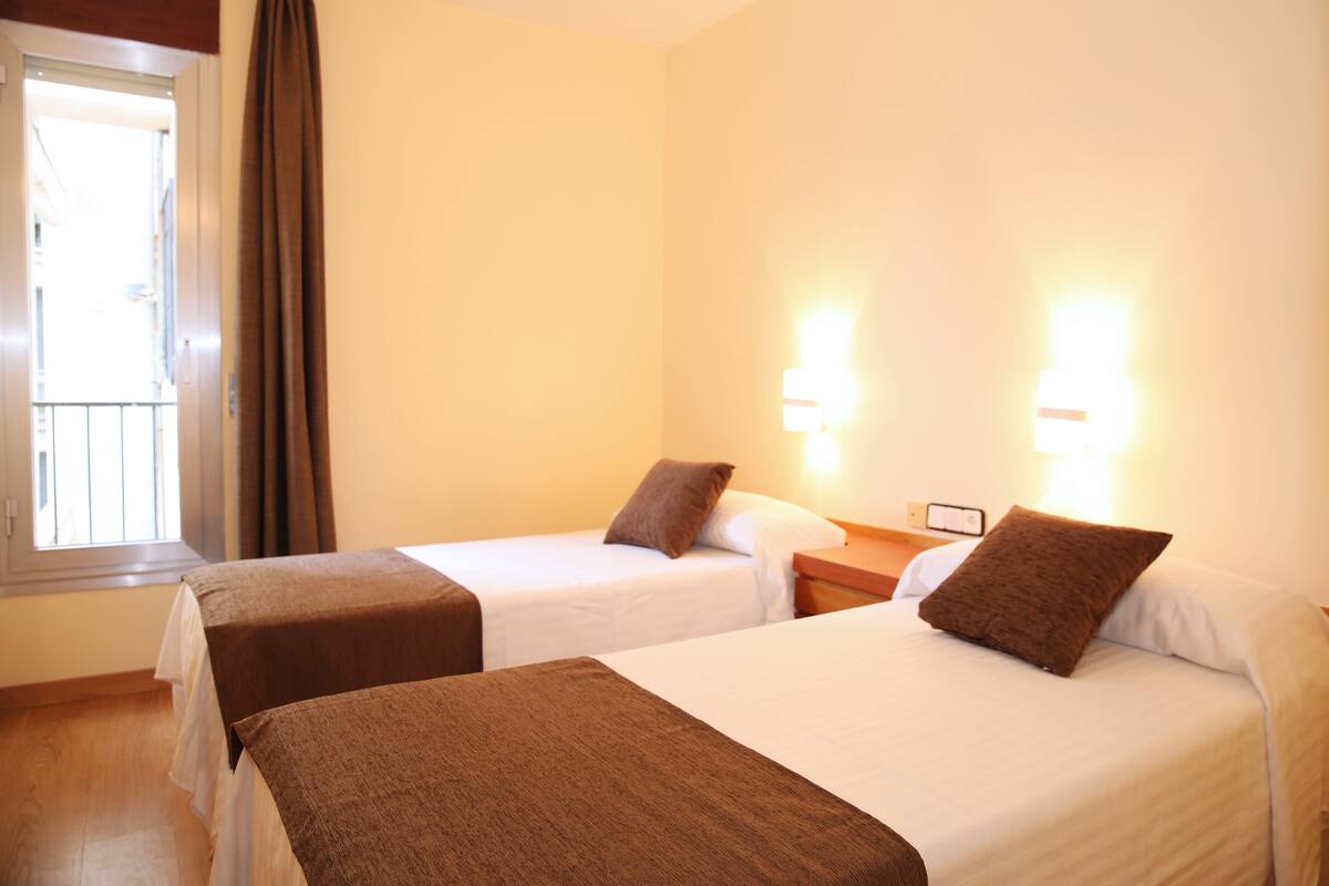 A hotel room with two beds, a window with curtains, and wall-mounted lights.