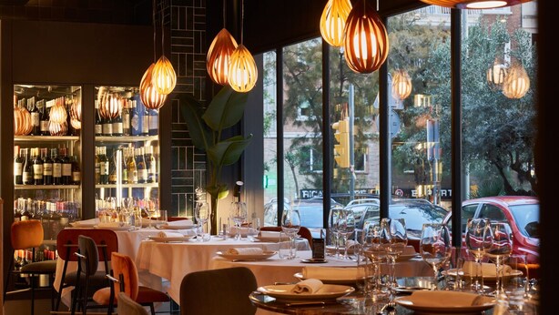 Un restaurante con mesas dispuestas para comer, amplias ventanas con vista a la calle y lámparas colgantes.