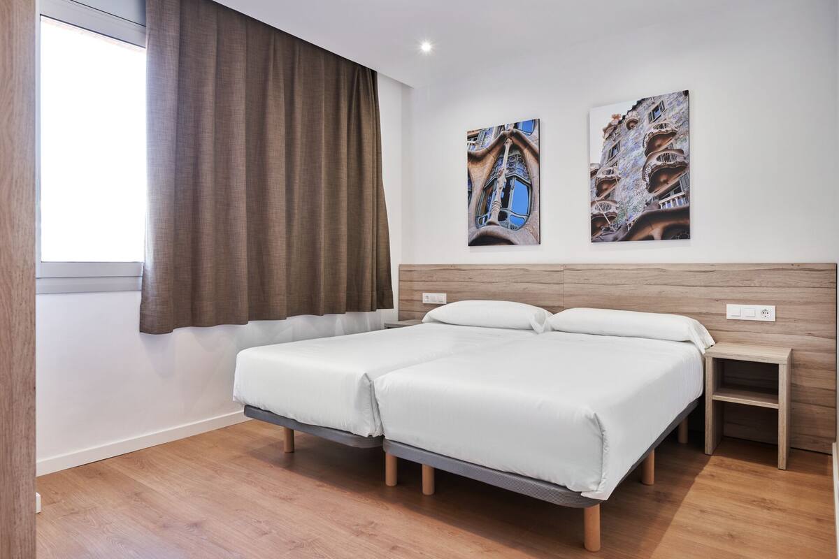 A hotel room with a double bed, a small bedside table, a window with curtains, and two wall-mounted artworks.