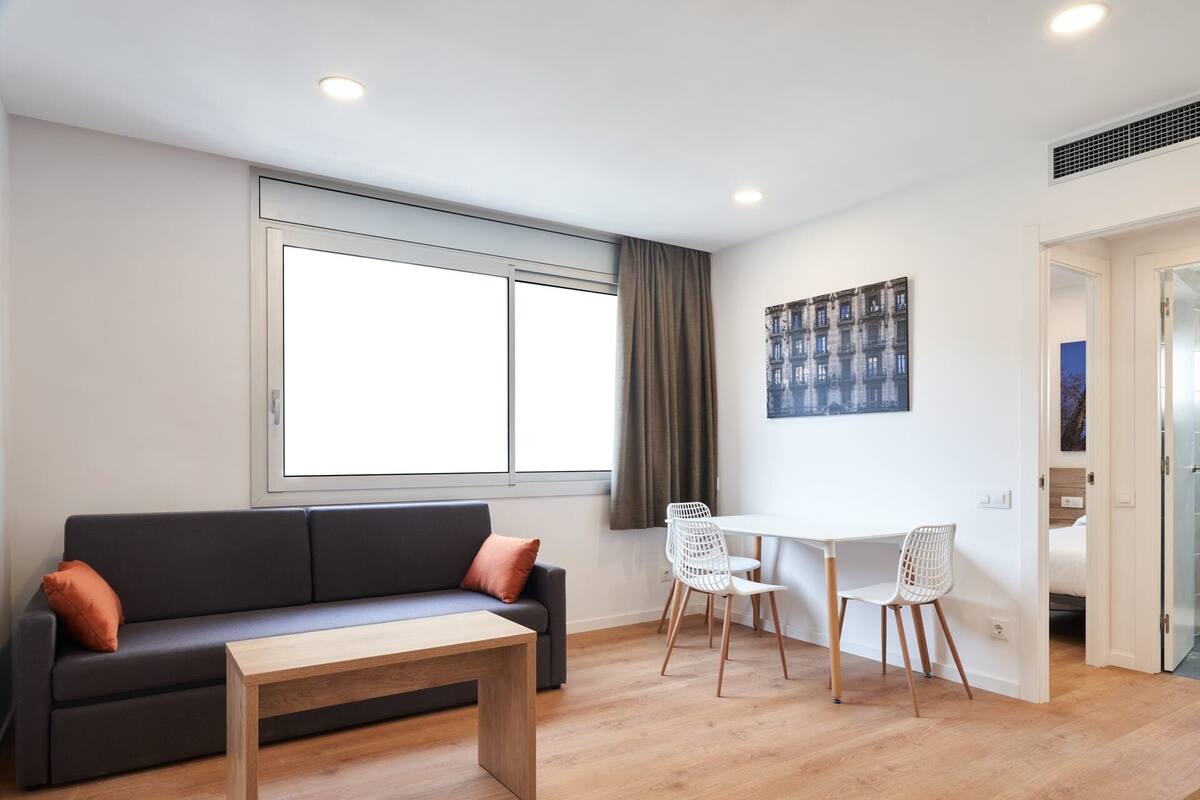 A modern living room with a dark grey sofa, a wooden coffee table, and a white dining table with two white chairs. There is a large window with a view of buildings, a painting on the wall, and a door leading to another room.