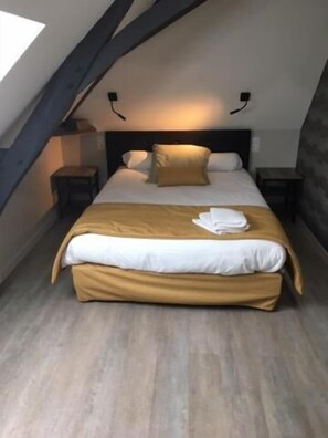 Une chambre avec un plafond en pente, un lit recouvert d’une couverture de couleur beige, des oreillers blancs et une serviette pliée posée sur le lit. Il y a deux tables de chevet avec des lampes.