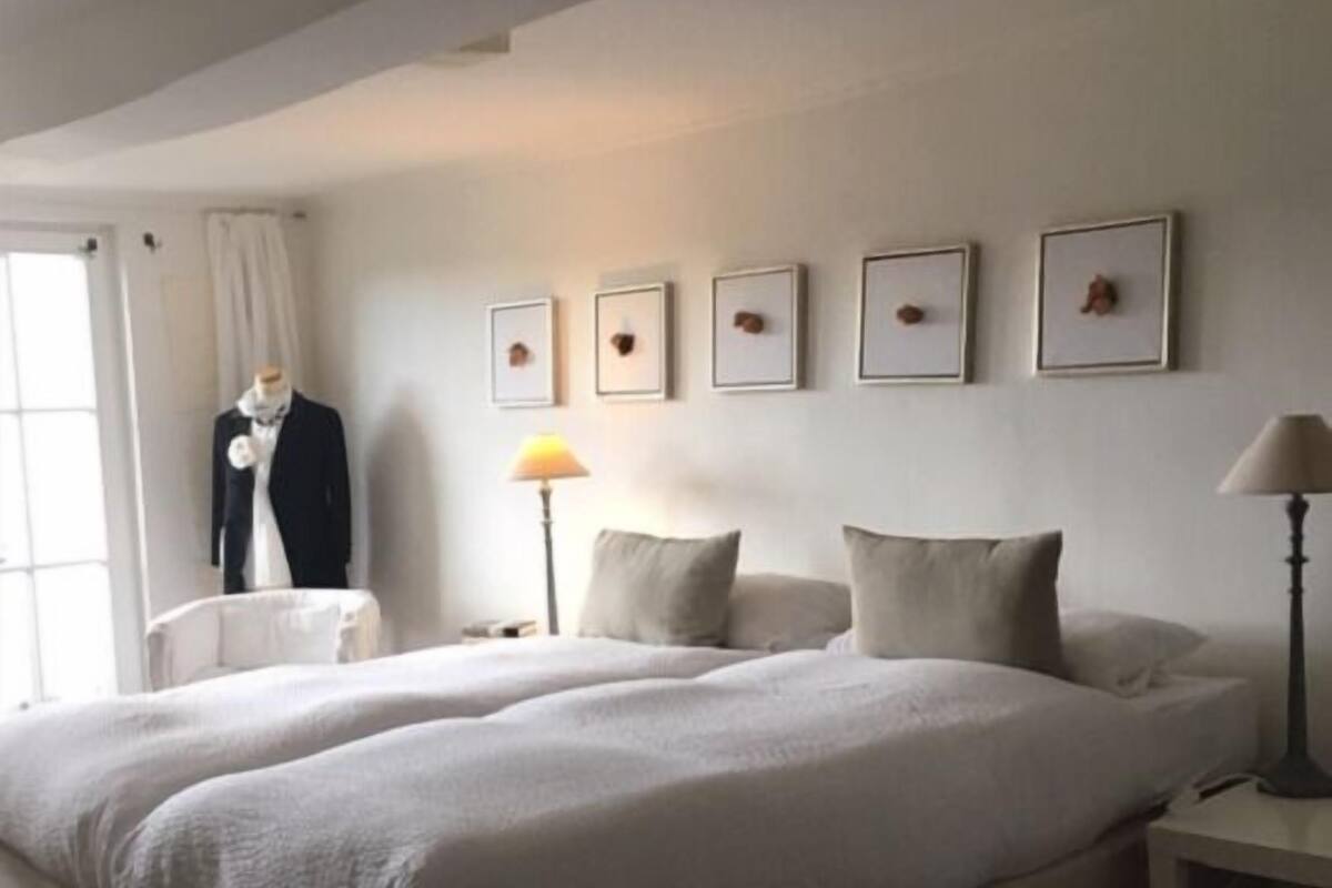 Ein Doppelbett mit einer neutralen Farbgestaltung, eine Büste in einem Anzug und gerahmte Bilder an der Wand.