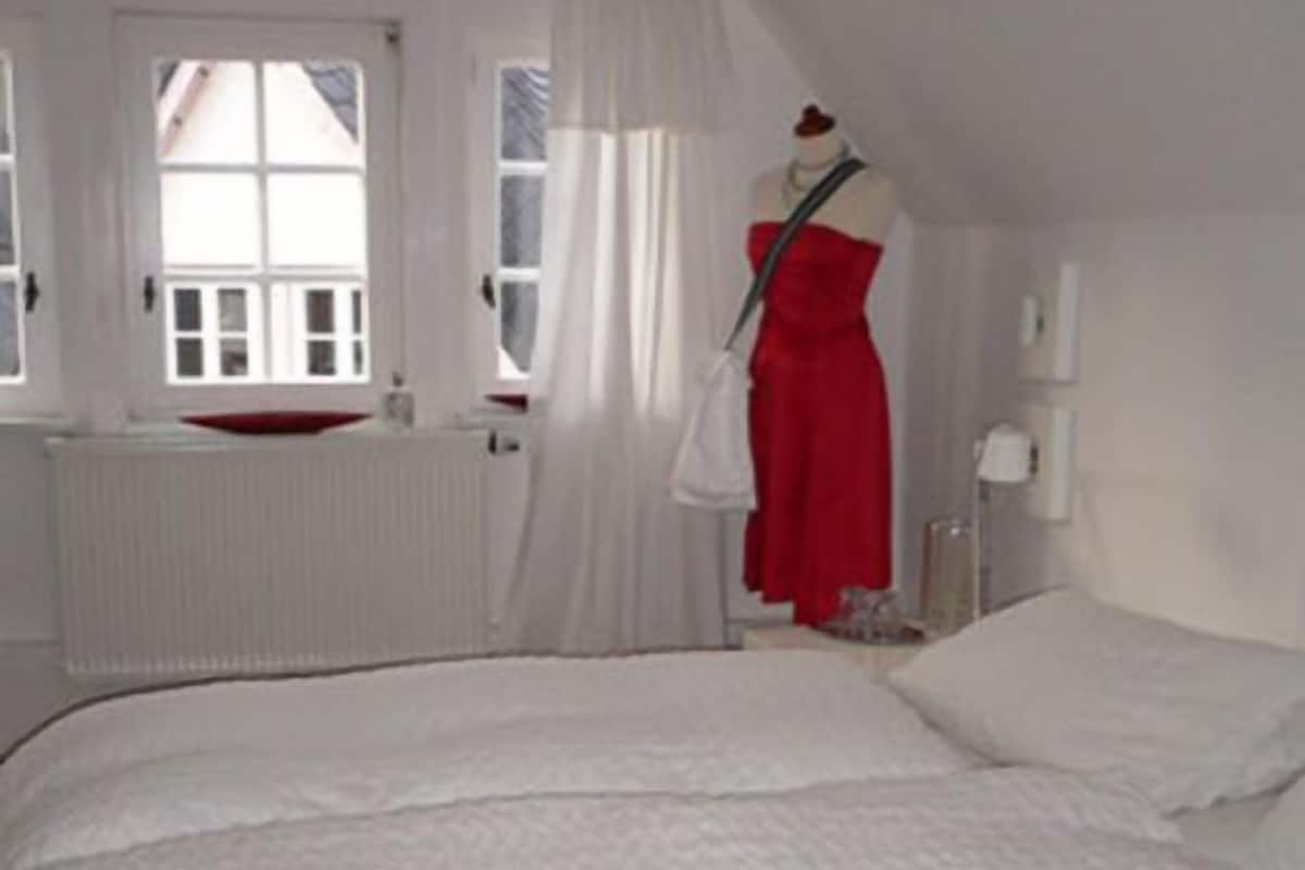 Ein Schlafzimmer mit einem großen Bett, einem weißen Heizkörper, einem Fenster mit weißen Vorhängen und einer Puppe in einem roten Kleid.