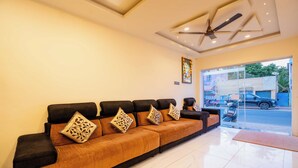 A hotel lobby with a long, upholstered sofa with patterned cushions, a ceiling fan, and a large window offering a view of the street. - Hotel NNP Grand (Rameshwaram)