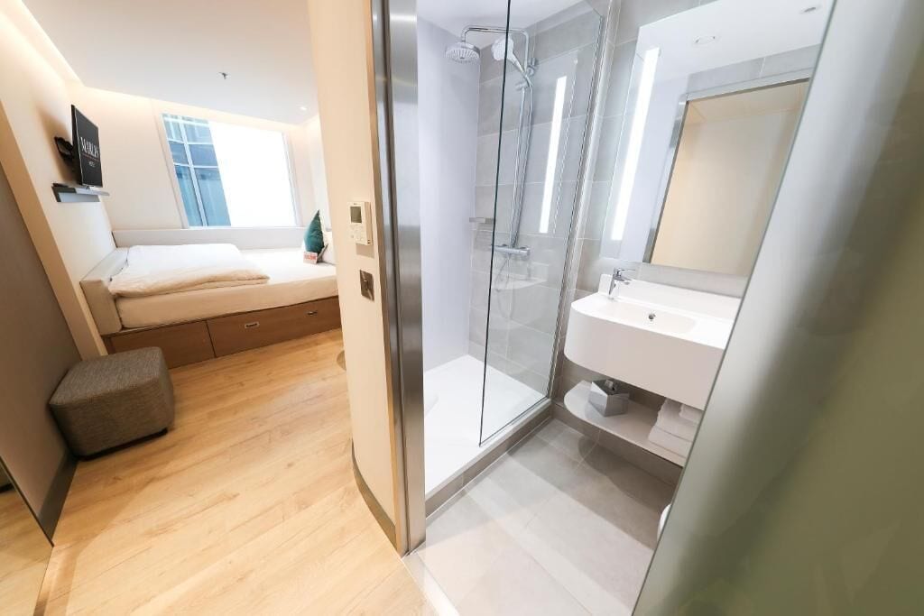 A modern hotel room with a bed, a glass shower enclosure, and a bathroom with a sink and mirror.