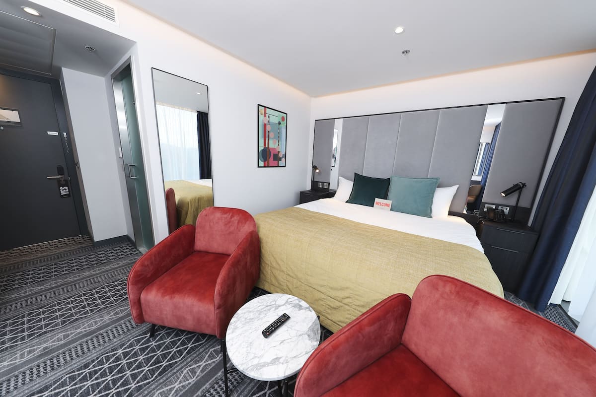 A hotel room with a large bed, two red armchairs, a small round table, and a large mirror.