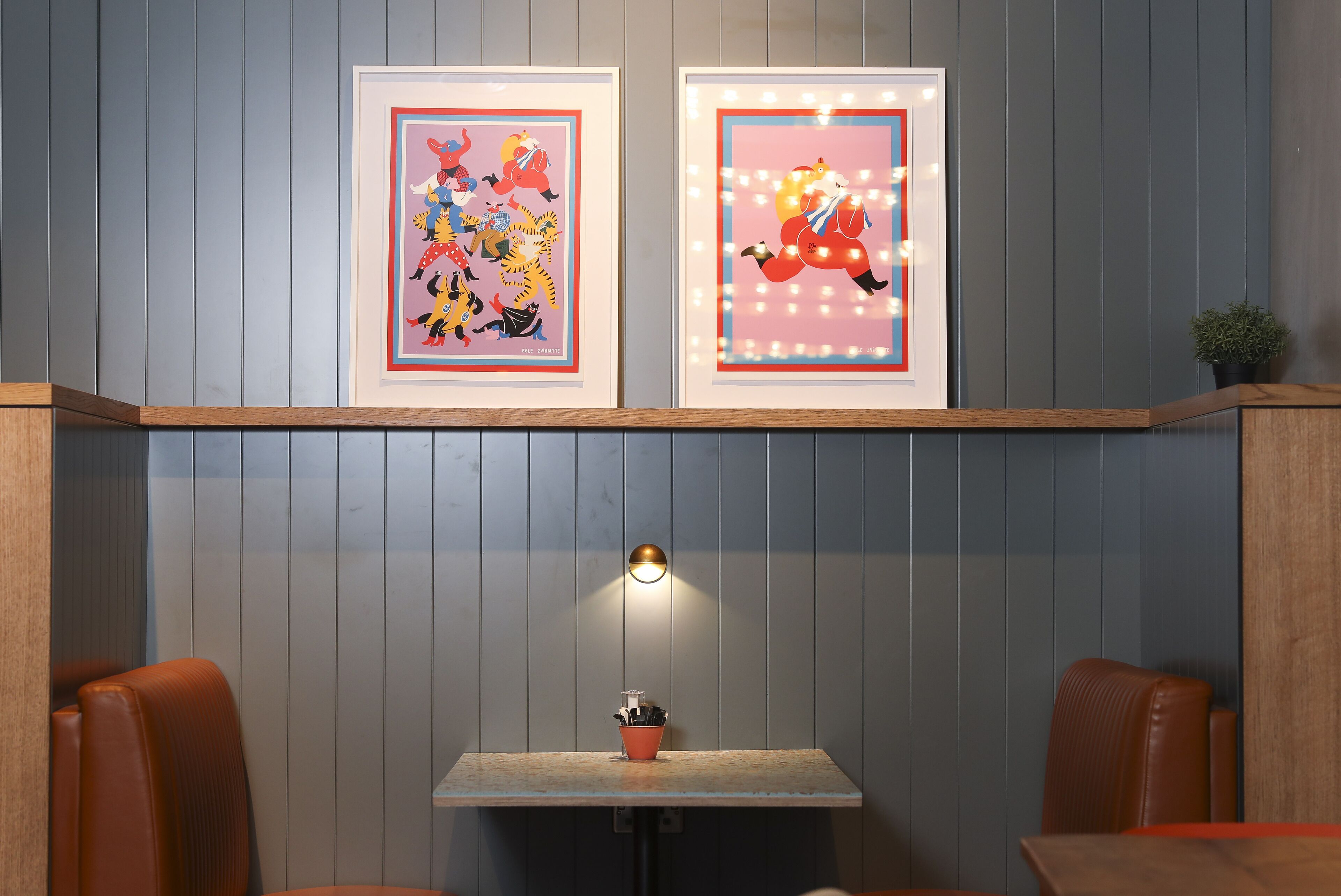 A restaurant with a wooden bench seating area, a small table with a red pot, and two framed artworks on the wall.