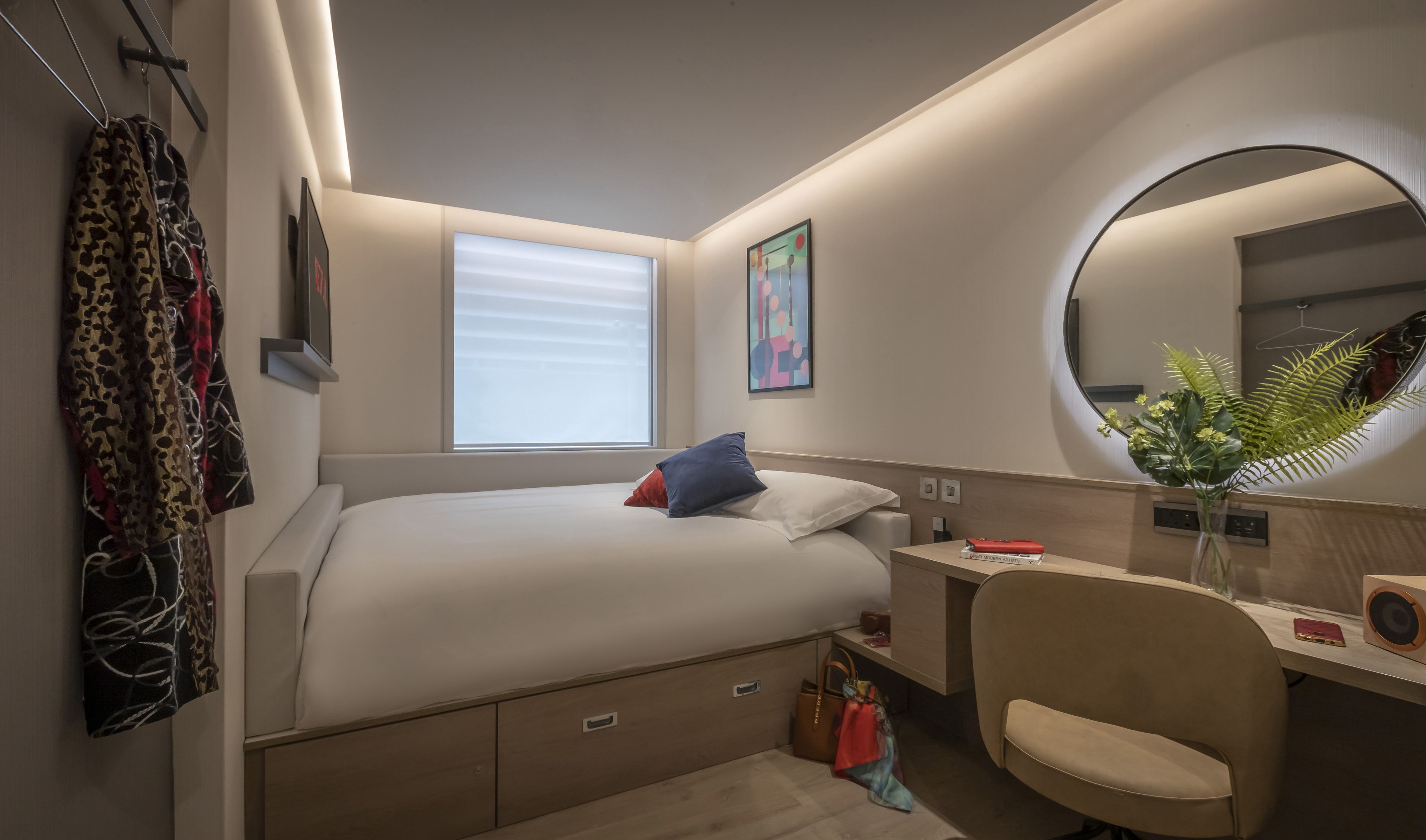 A modern hotel room with a bed, bedside table, chair, and a round mirror.