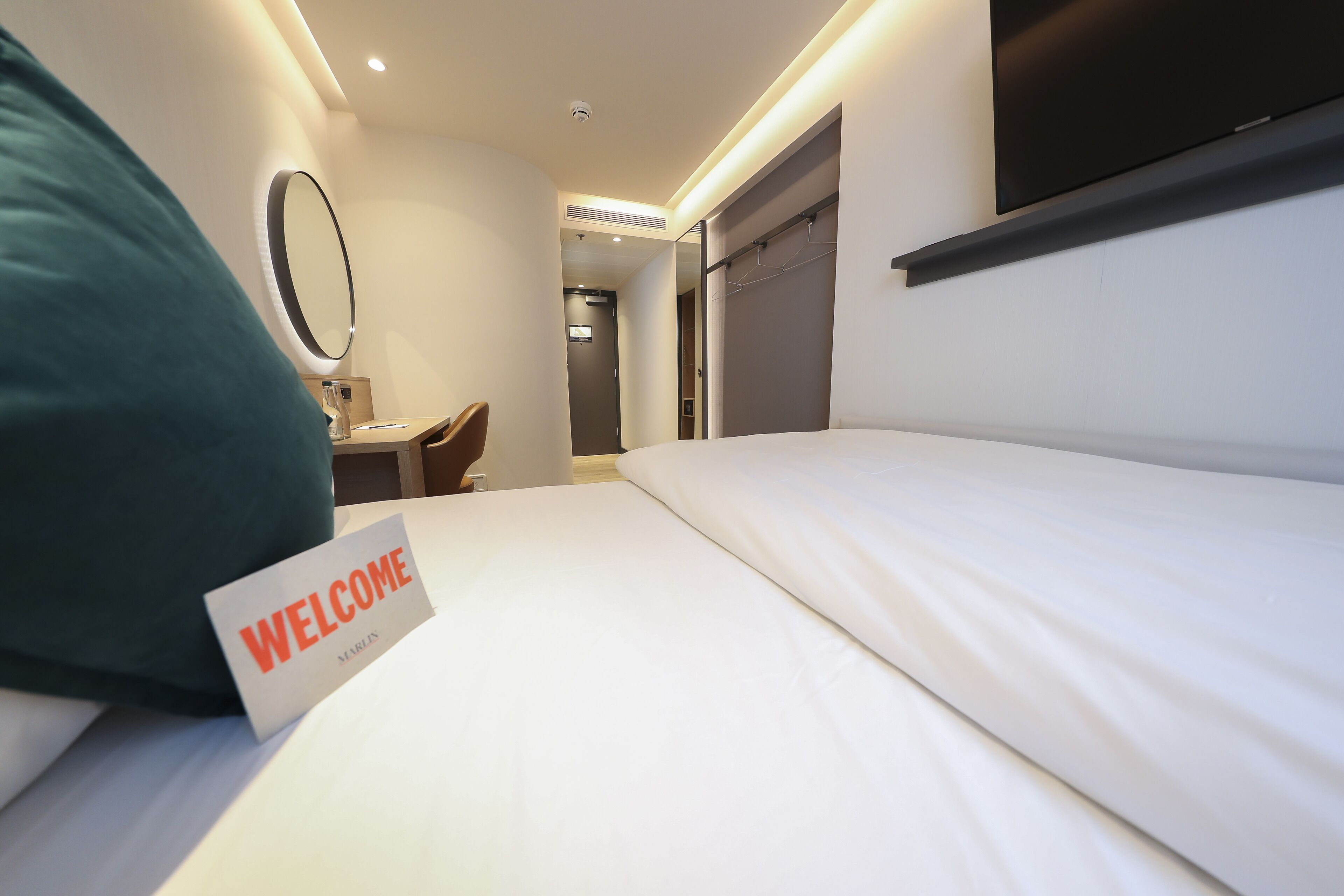 A hotel room with a bed, a 'WELCOME' sign, a mirror, a TV, and a door leading to a hallway.