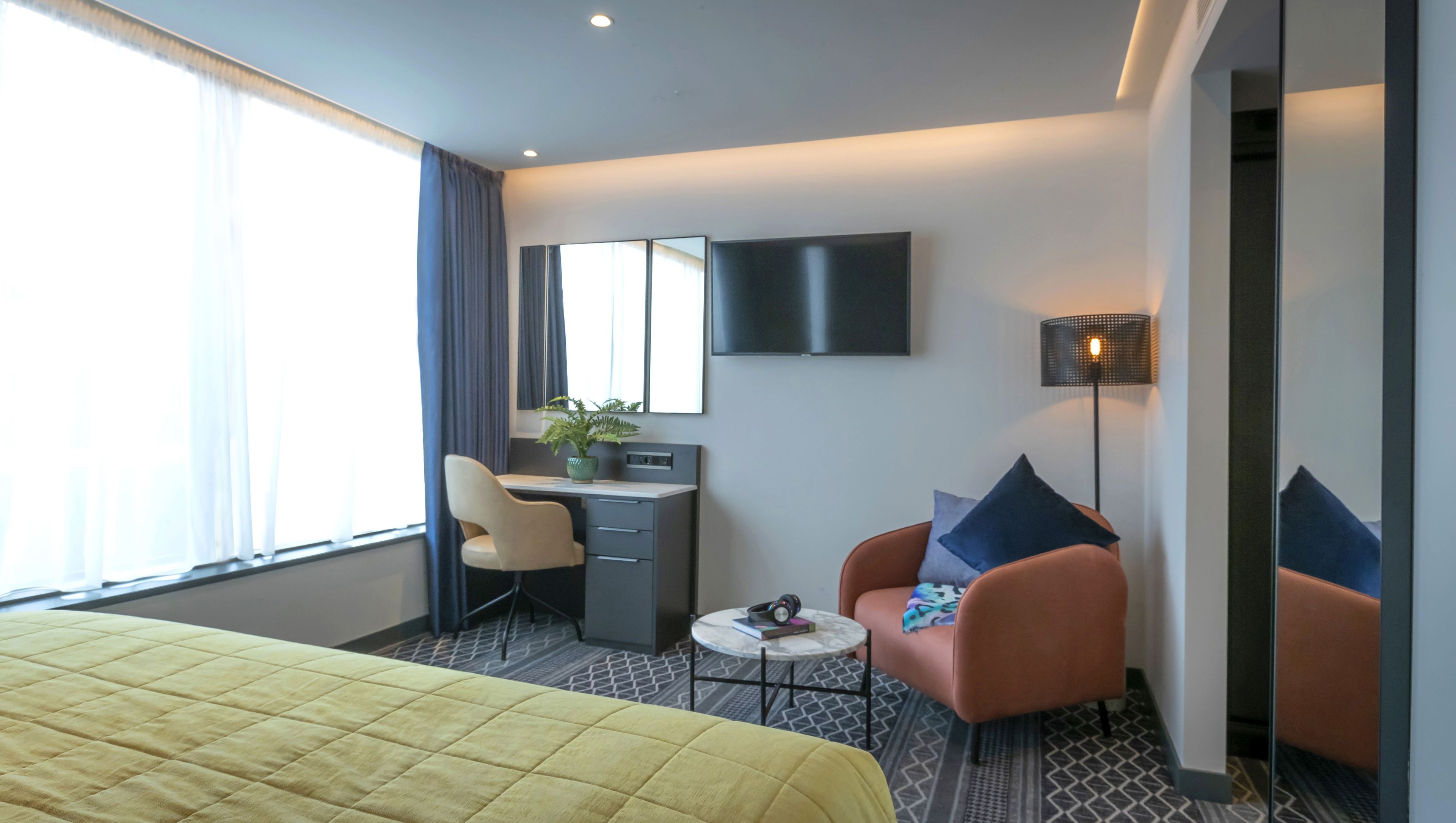 A hotel room with a bed, a desk, a chair, a sofa, a TV, and a lamp.