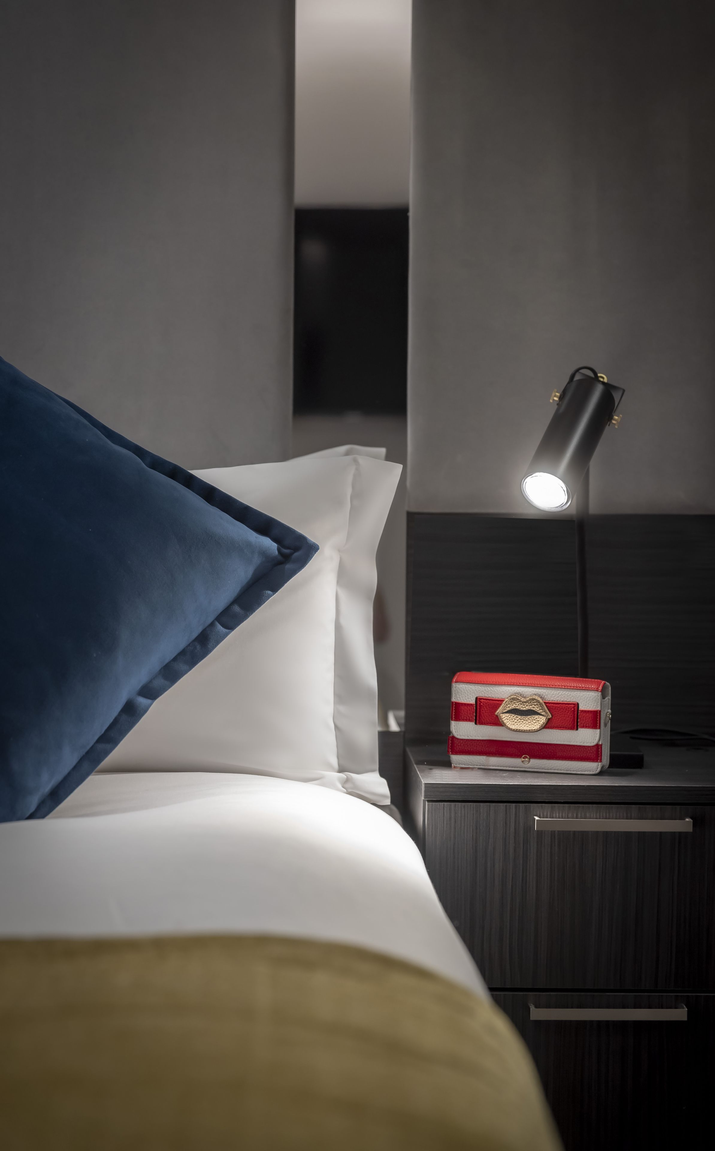 A neatly made bed with blue and white pillows, a bedside table with a lamp, and a red and white box on the table.