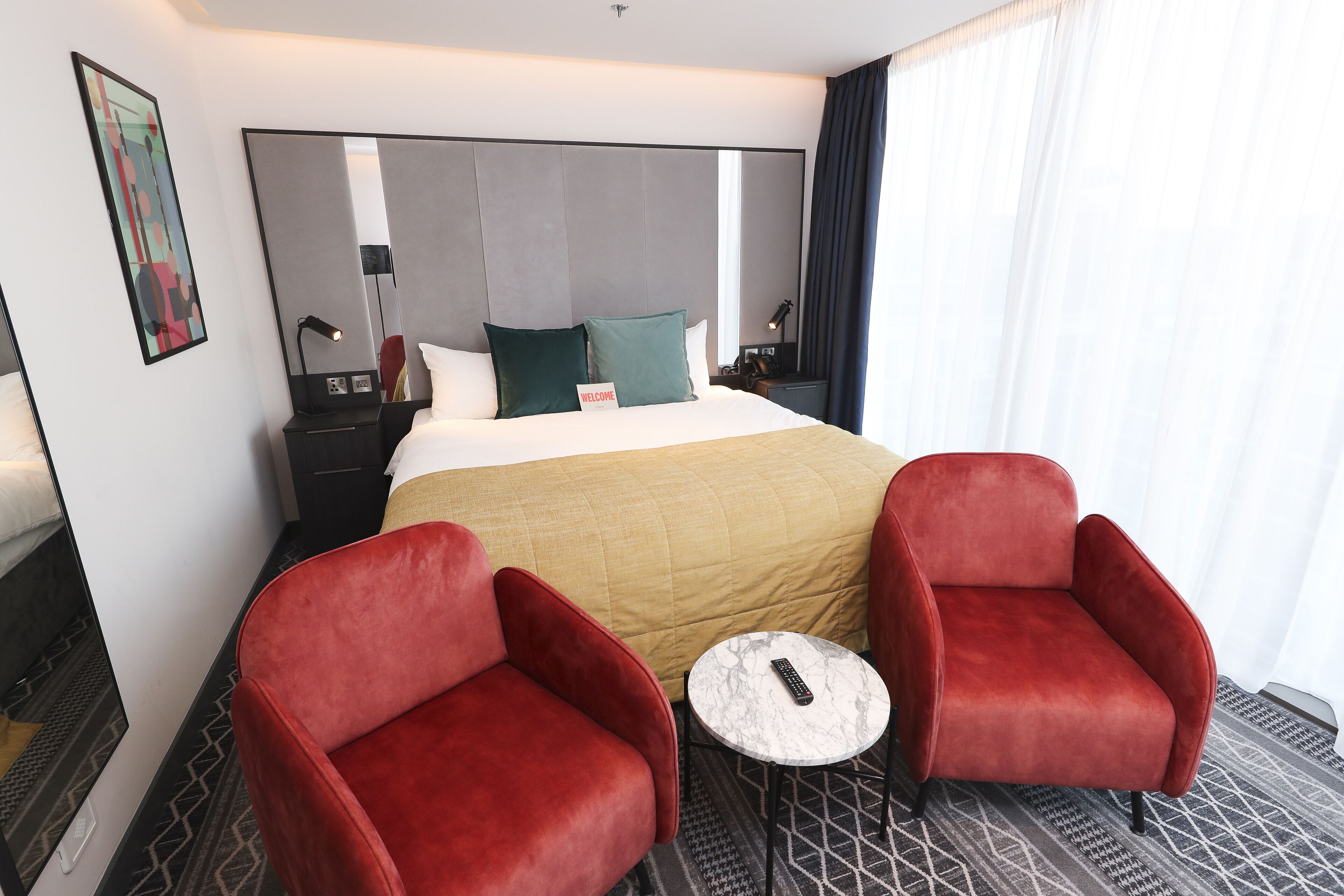 Une chambre d’hôtel avec un grand lit, deux fauteuils rouges, une petite table ronde et une table de chevet avec une lampe.