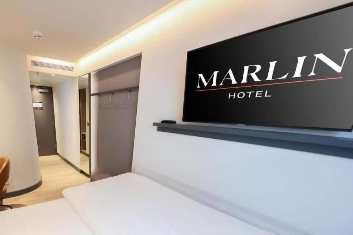 A modern hotel room with a bed, a desk, a chair, and a large mirror. The Marlin Hotel sign is visible on the wall.