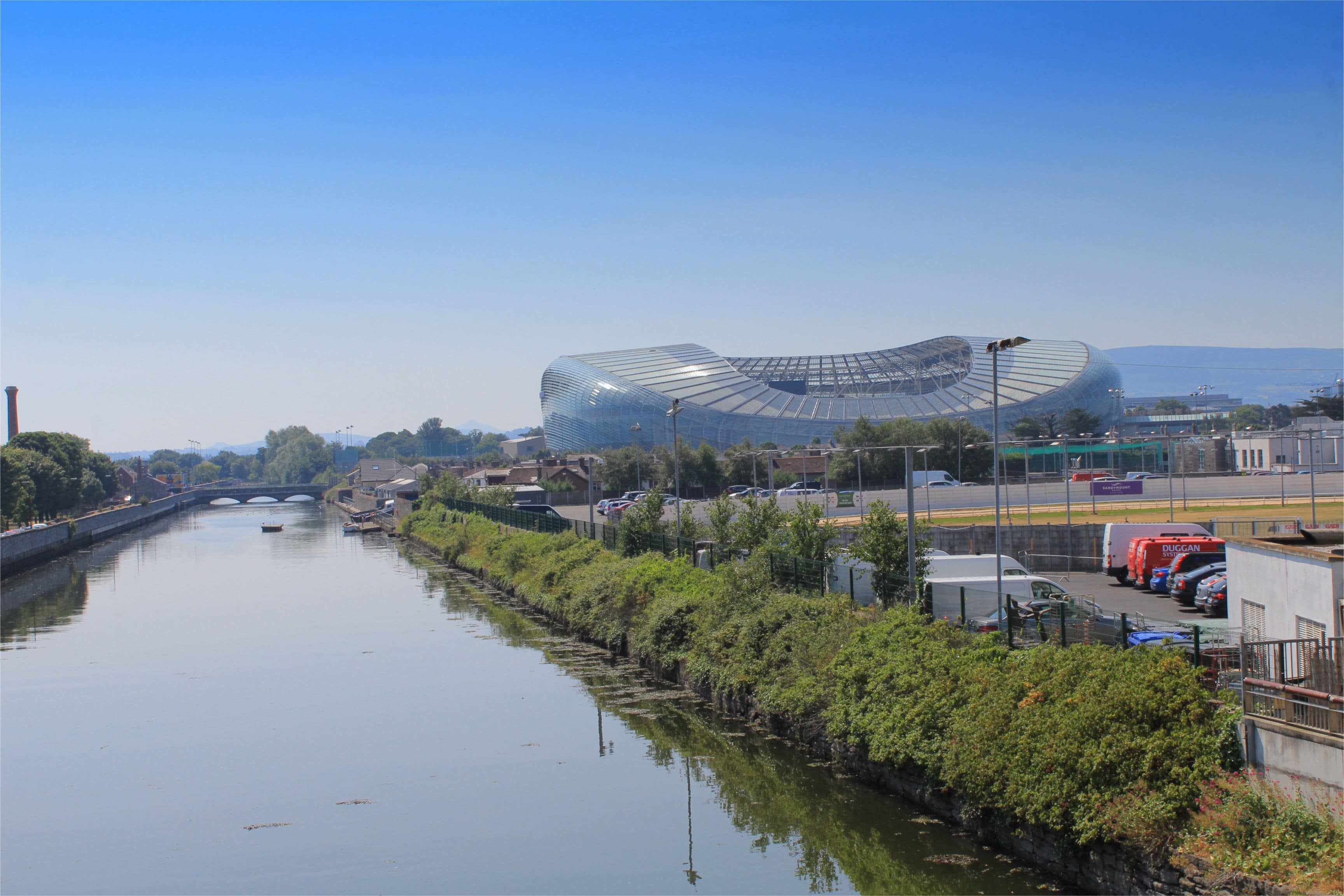A modern building with a unique curved design, a river running alongside, and a bridge in the background.