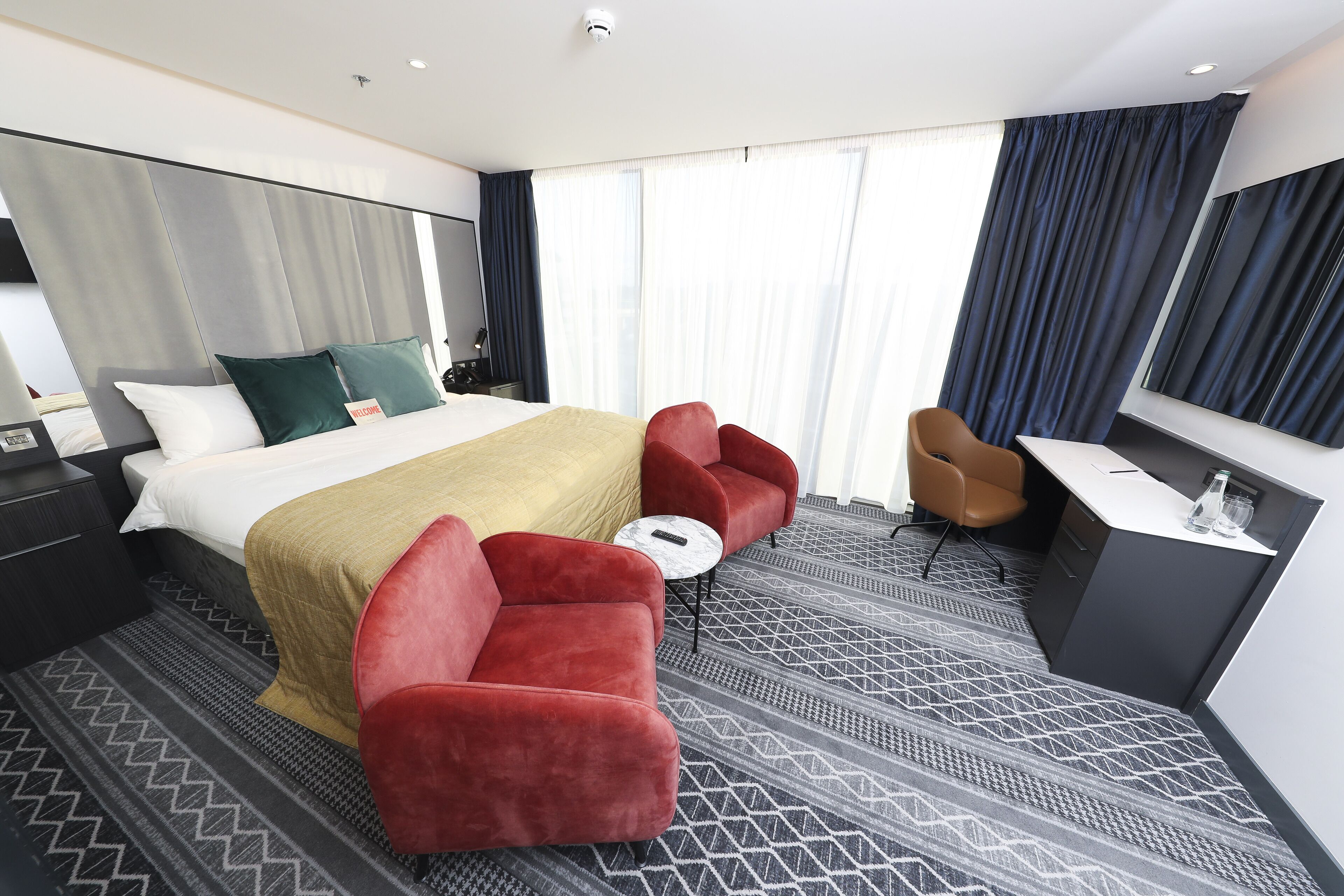A modern hotel room with a large bed, red armchairs, a small round table, a desk, and a large window with curtains.