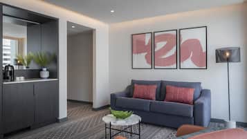 Una sala de estar moderna con un sofá gris, una mesa de centro y una zona de cocina con un mueble oscuro y una planta.