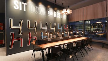 A modern seating area with a long table and chairs, a wall with illuminated letters, and a view of the street outside.