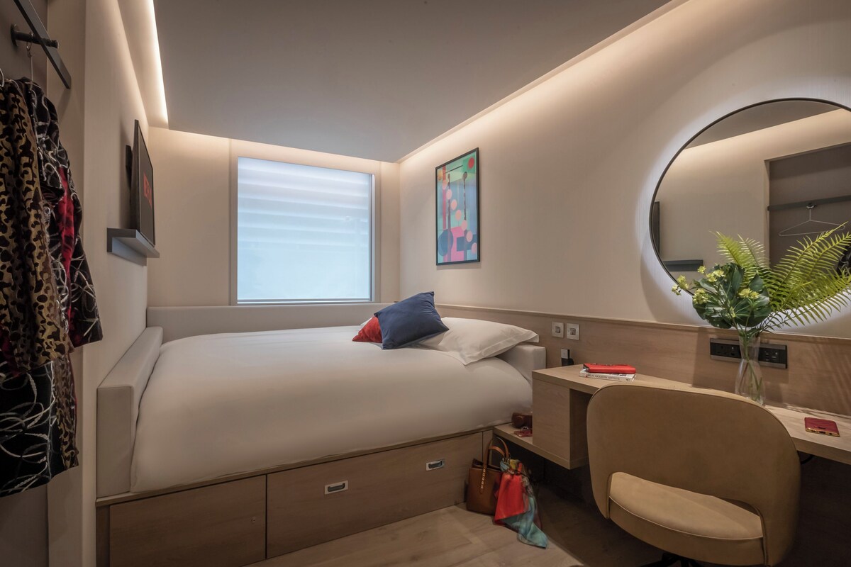 A modern hotel room with a bed, bedside table, chair, and a round mirror.