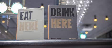 Two signs on a counter reading "EAT HERE" and "DRINK HERE" with a metal rack of glasses above them.