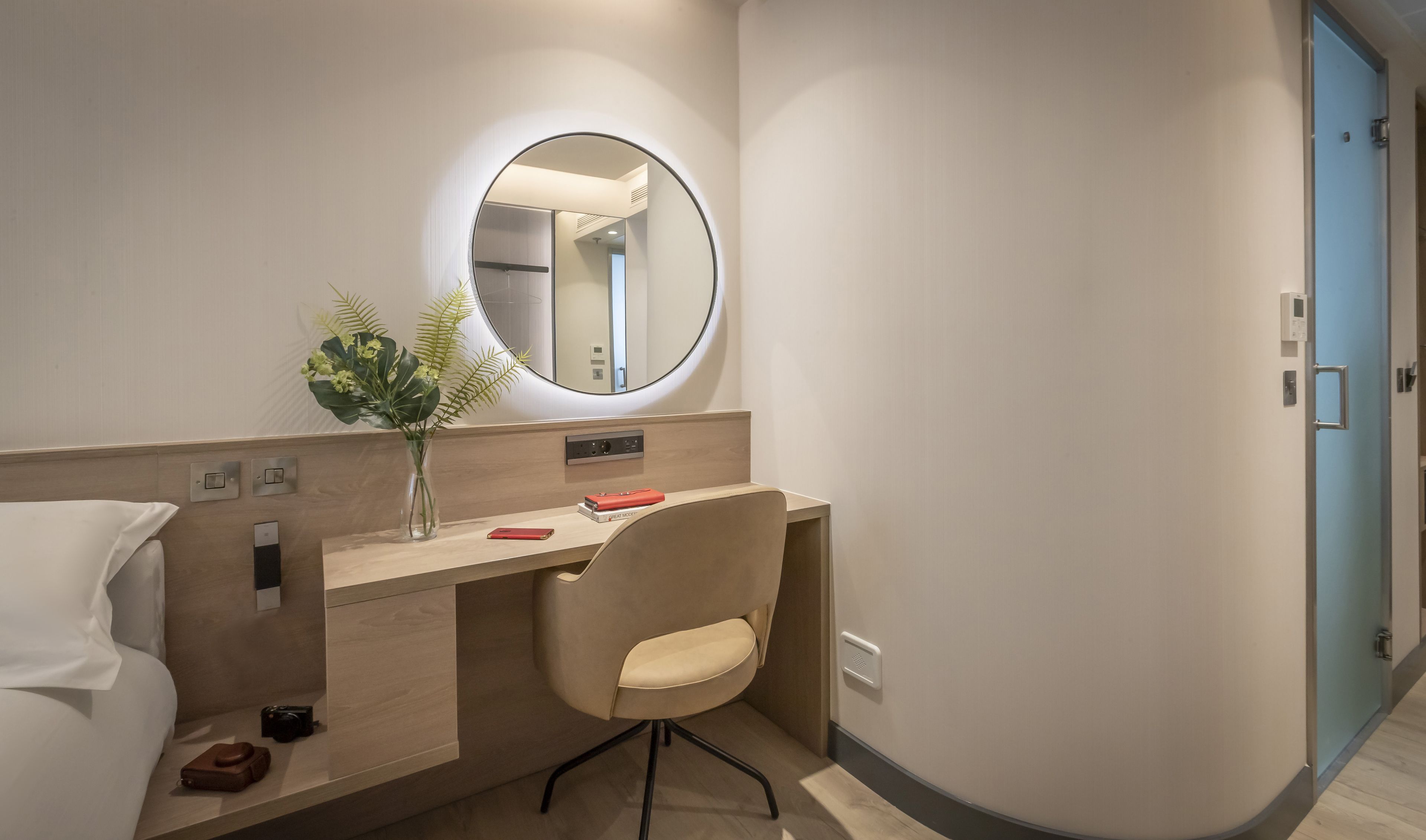 A modern hotel room with a wooden desk, a chair, a round mirror, and a vase with plants.