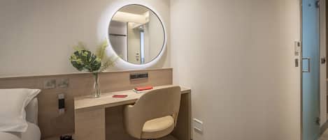 A modern hotel room with a wooden desk, a chair, a round mirror, and a vase with plants.