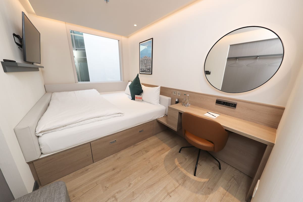 A modern hotel room with a bed, desk, chair, and a large mirror.
