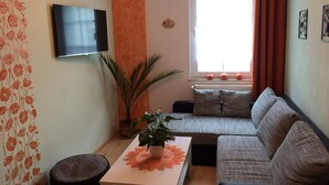 A living room with a sofa, a TV, a window with curtains, a plant, and a table with a placemat. - Pension Zum Bauernstübl (Meerane)