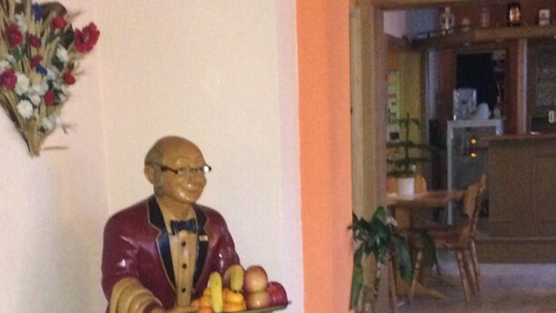 A statue of a man holding a tray with fruits and vegetables in a lobby area with a potted plant and a dining table in the background. - Pension Zum Bauernstübl (Meerane)