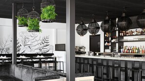 Un área de bar moderna con taburetes negros, una barra con una variedad de botellas y plantas verdes colgantes en cestas de alambre.
