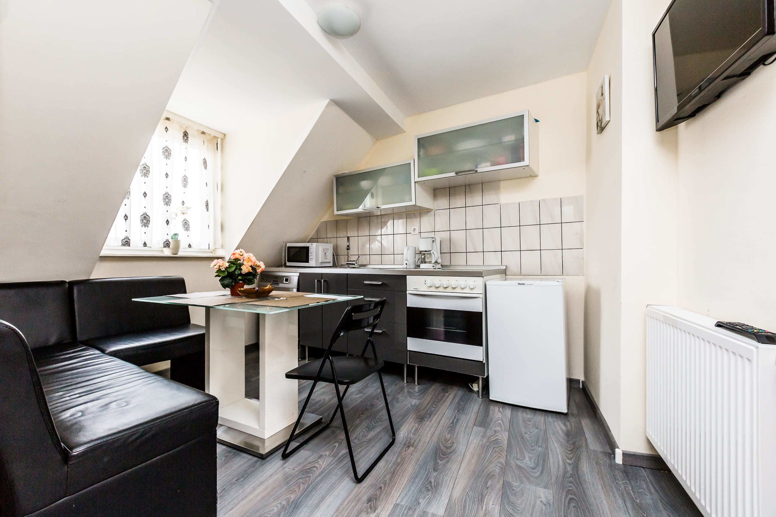 Eine kompakte Küche mit Essbereich, einem schwarzen Ledersofa, einem kleinen Tisch mit einer Vase und einem Fenster mit gemusterten Vorhängen.