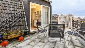 A small, compact house with a tiled roof, a sliding glass door leading to a kitchen, and a patio area with metal chairs and a wooden cart. - Tripcologne Apartments since 2013 (Cologne)