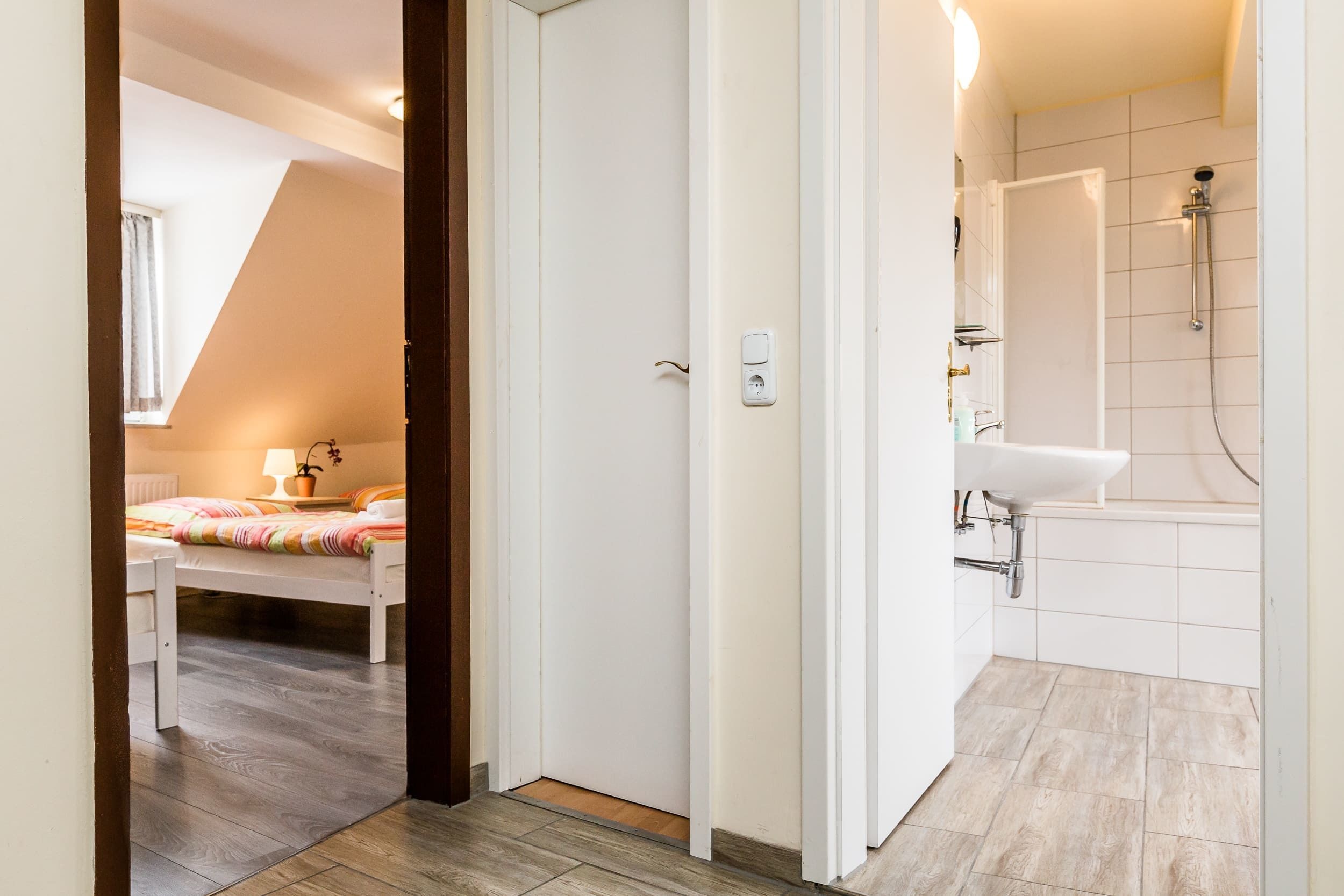 Ein Schlafzimmer mit Bett, Nachttisch und Fenster. Daneben ein Badezimmer mit Dusche, Waschbecken und gefliesten Wänden.