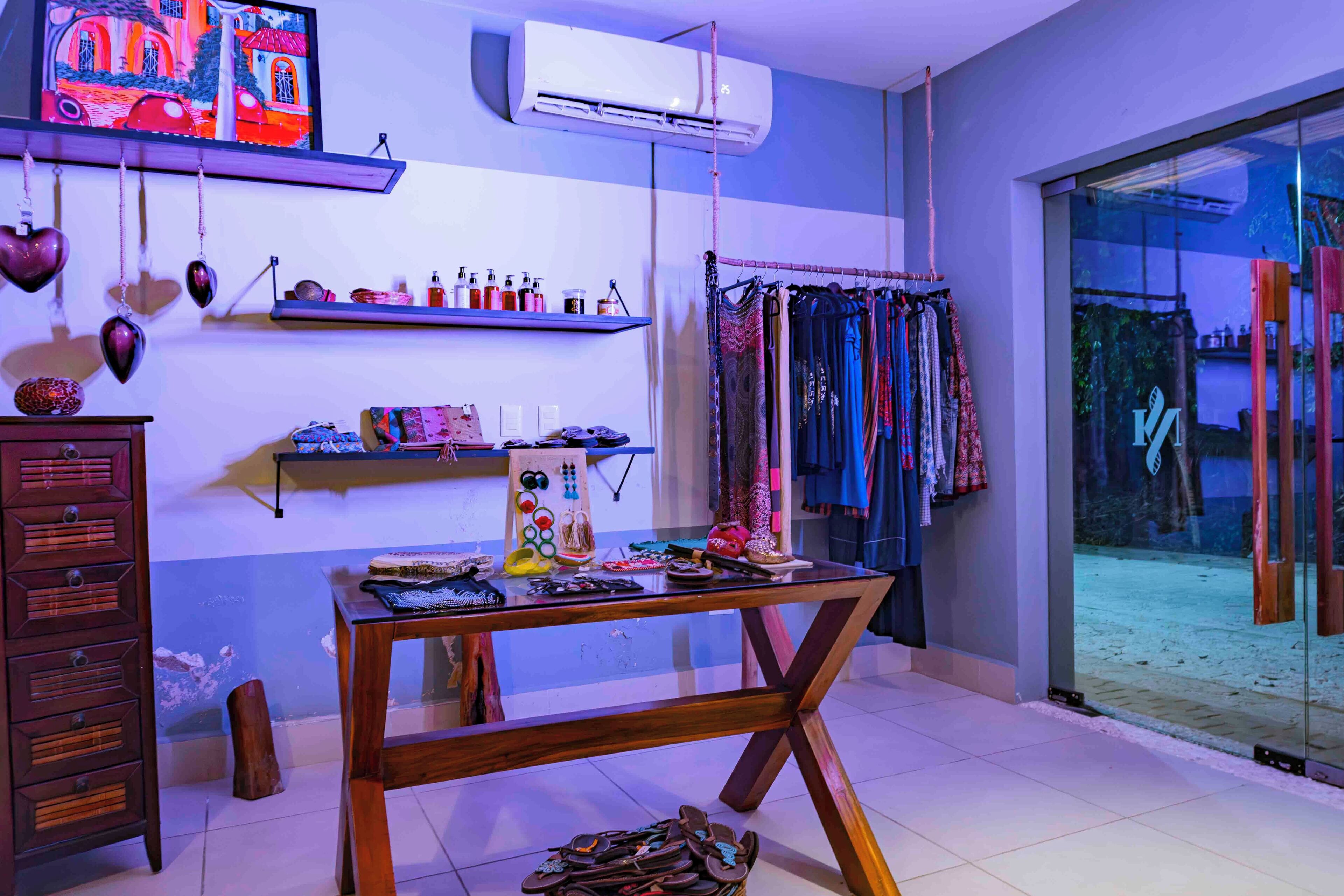 A room with a wooden table displaying various items, a dresser, shelves with products, and a view through a glass door.