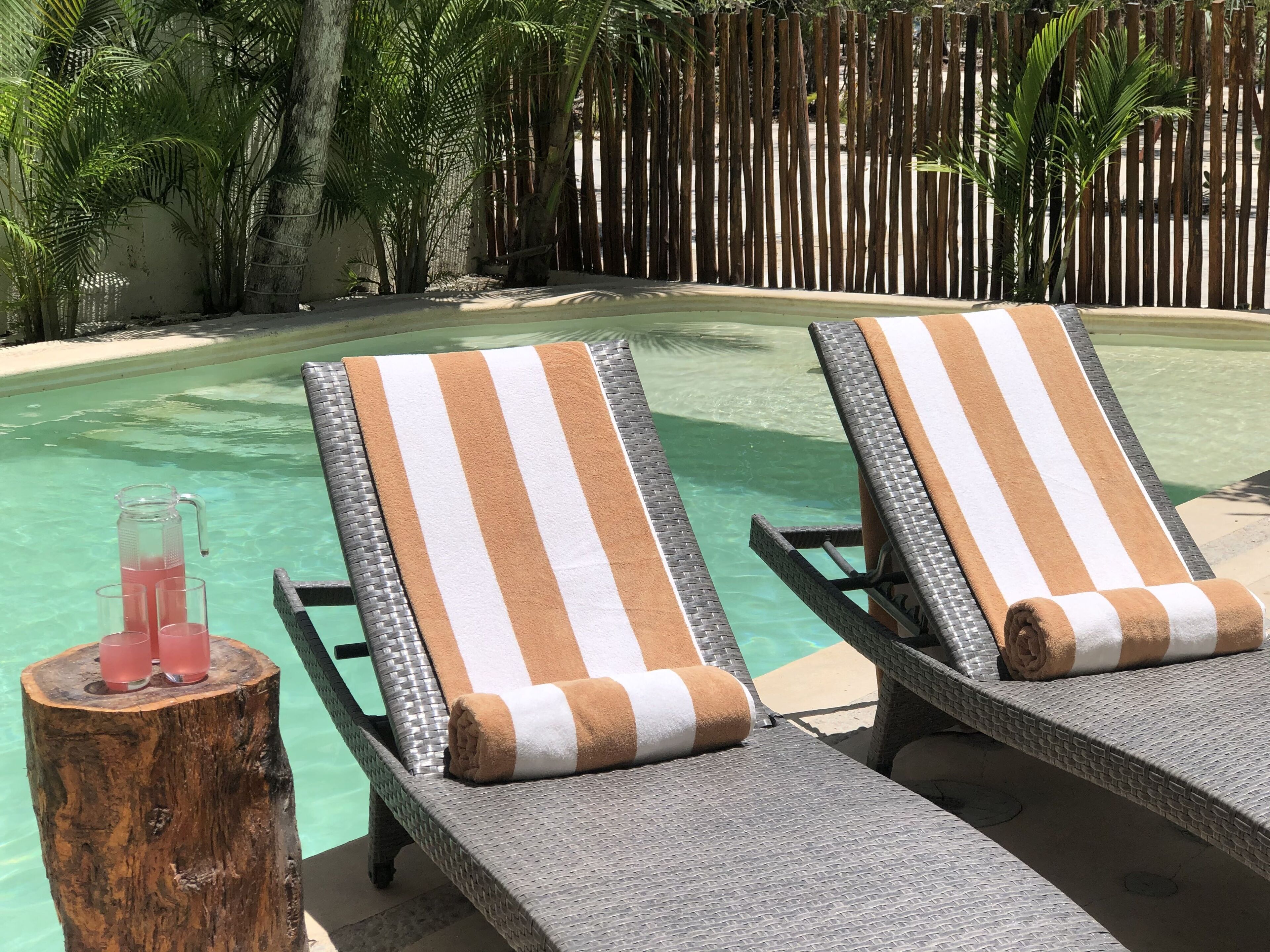 Un espace aménagé au bord de la piscine, avec deux chaises longues, un tronc d’arbre servant de table sur lequel est posée une carafe en verre, et une clôture en bois en arrière-plan.