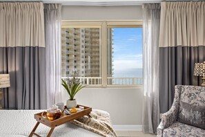 Een hotelkamer met een bed, een houten dienblad met thee en gebak, een potplant, een stoel, en een balkon met uitzicht op een hoog gebouw en de zee.