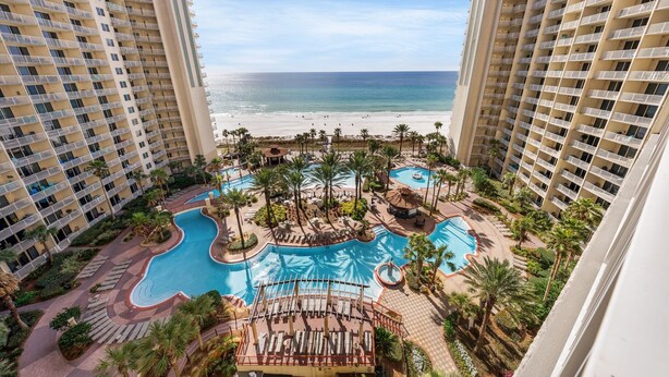 A high-rise hotel with a pool area, palm trees, and a beach view.