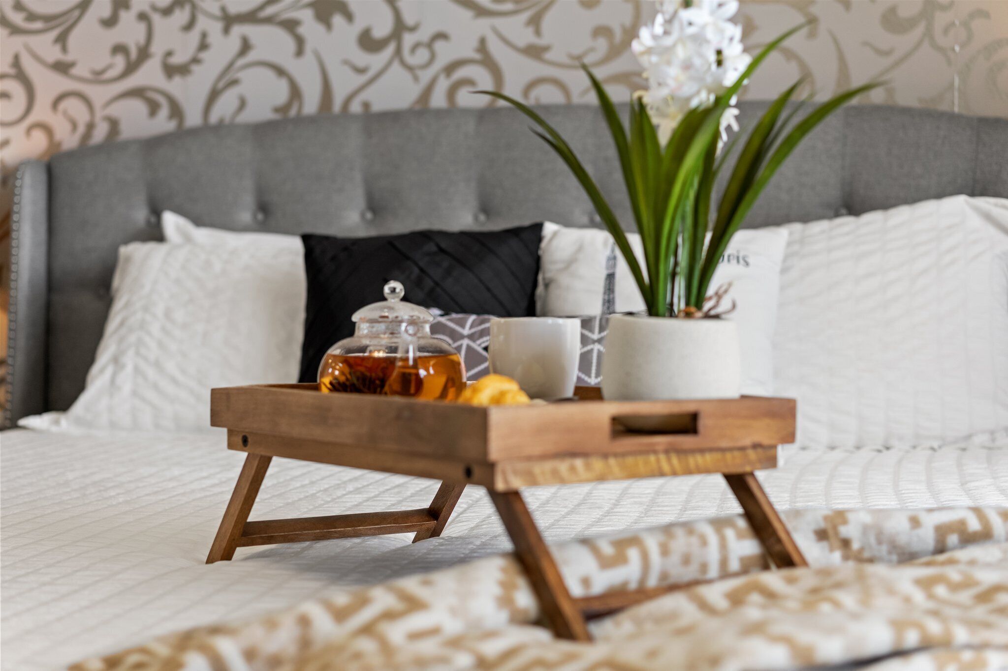 A neatly made bed with a grey headboard, white pillows, and a tray with a glass jar, a cup, and a plant.
