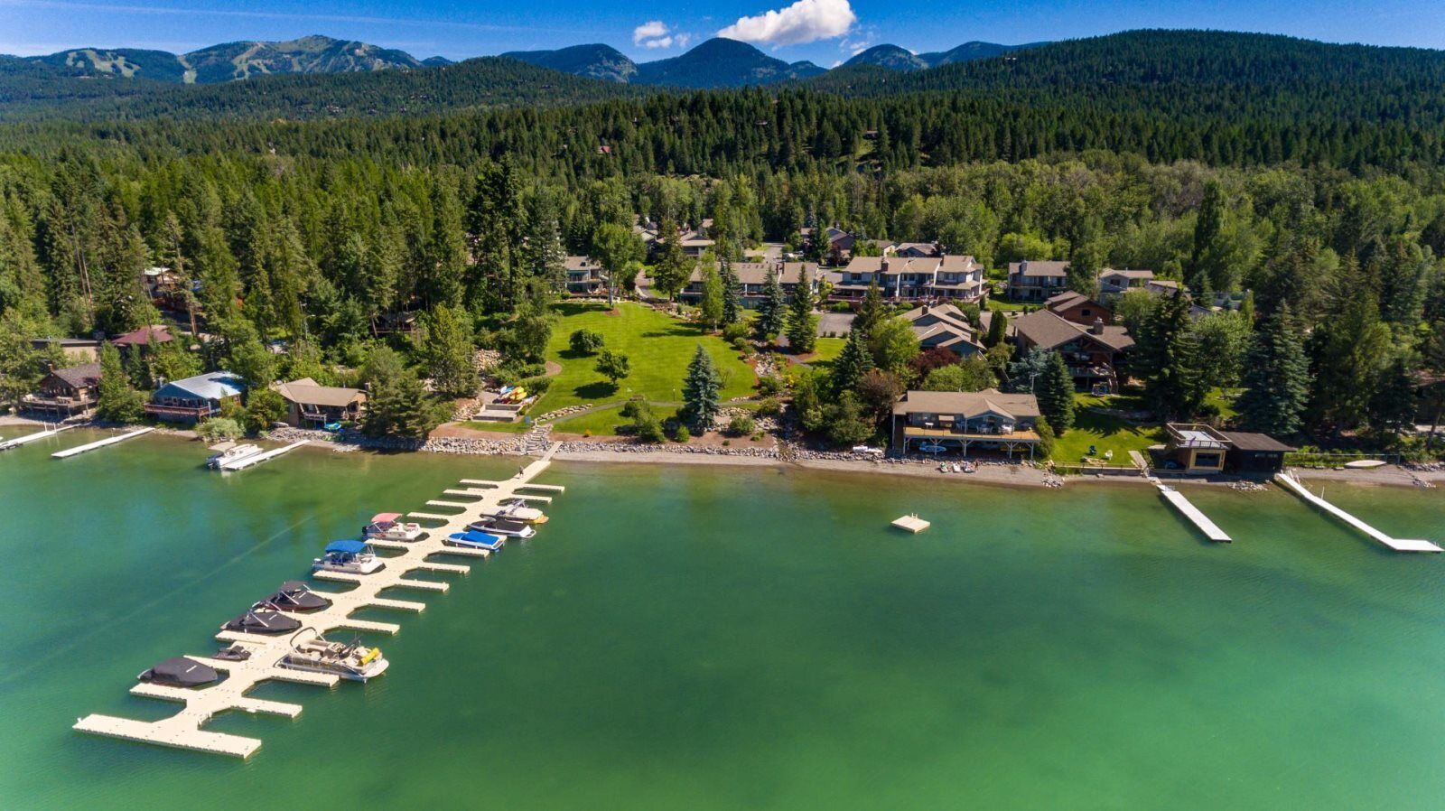 Vista aérea de un complejo turístico junto a un lago, con un puerto deportivo, casas y un paisaje boscoso de fondo.