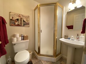 A bathroom with a white toilet, a glass-enclosed shower, a sink with toiletries, and a poster on the wall.