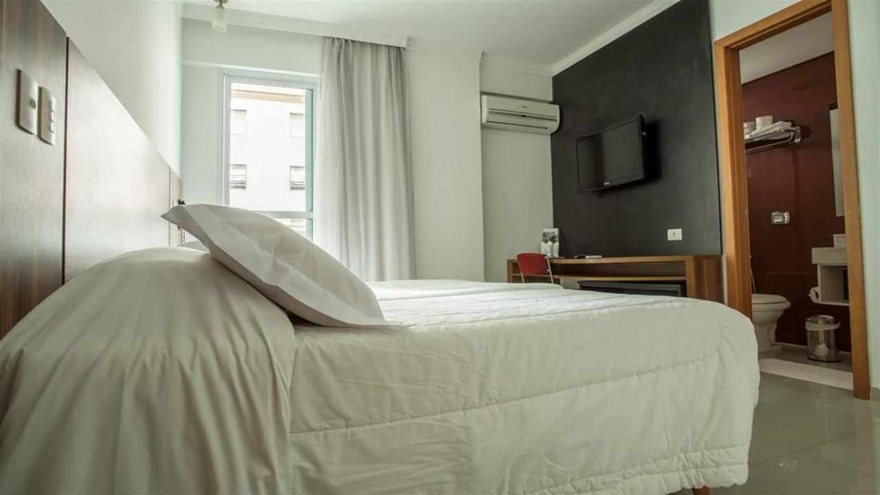 A hotel room with a bed, a television mounted on the wall, an air conditioning unit, a window with curtains, and a bathroom with a toilet and sink.