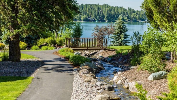A pathway leading to a wooden bridge over a stream, surrounded by greenery and a lake in the background. - Mountain Harbor #137 3 Bedroom Condo (Whitefish)