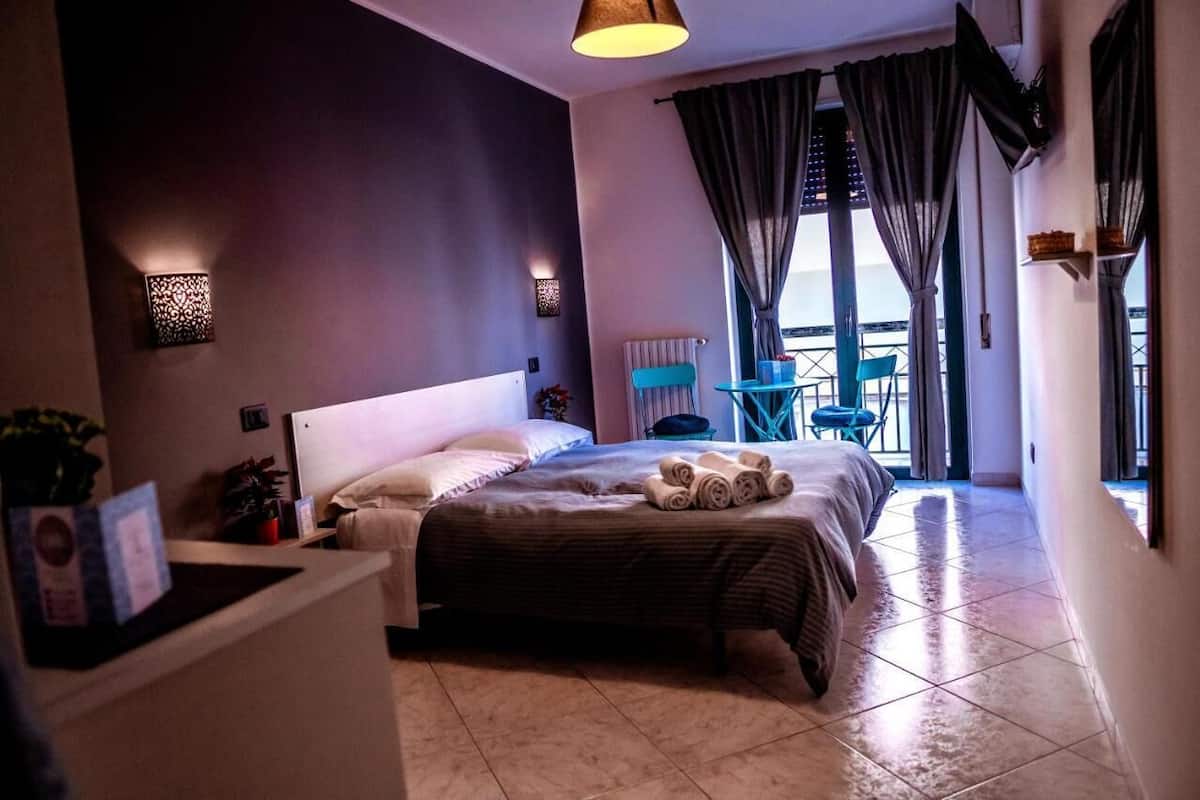 A hotel room with a large bed, a small table, a chair, and a balcony with a view.